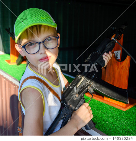 portrait of a boy in glasses and a bright green cap with a black automatic rifle in his hands near range stand portrait of a boy in glasses and a bright green cap with a black automatic rifle in his hands near range stand 140580284