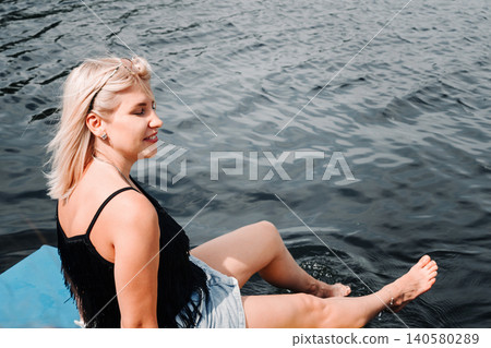 blonde girl sitting on the edge of a blue pier by the water with her eyes closed blonde girl sitting on the edge of a blue pier by the water with her eyes closed 140580289