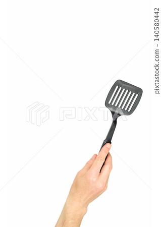 a human hand raises up a black plastic slotted spatula on a white background a human hand raises up a black plastic slotted spatula on a white background 140580442