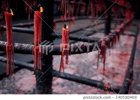 Red candles burning on a metal holder with wax drippings, creating a serene atmosphere in a dimly lit space, showcasing the beauty of candlelight Red candles burning on a metal holder with wax drippings, creating a serene atmosphere in a dimly lit space, showcasing the beauty of candlelight 140580469