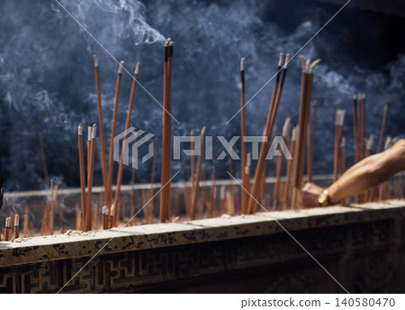 Incense sticks burning in a decorative holder with smoke rising, hand of a person visible, creating a serene atmosphere in a spiritual setting Incense sticks burning in a decorative holder with smoke rising, hand of a person visible, creating a serene atmosphere in a spiritual setting 140580470