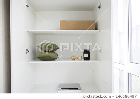 Modern white shelving unit with green decorative pillows, a black candle, an hourglass, and a cardboard box, positioned near a window with natural light Modern white shelving unit with green decorative pillows, a black candle, an hourglass, and a cardboard box, positioned near a window with natural light 140580497