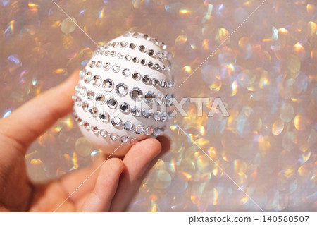 Decorative white Easter egg embellished with rhinestones held in a hand against a shimmering, blurred background of soft pastel colors Decorative white Easter egg embellished with rhinestones held in a hand against a shimmering, blurred background of soft pastel colors 140580507