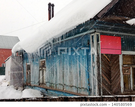 wooden blue house in winter during heavy snowfall under a roof with icicles wooden blue house in winter during heavy snowfall under a roof with icicles 140580569