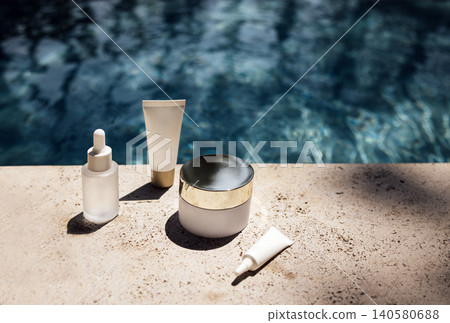 Skincare products arranged on a stone surface beside a swimming pool, featuring a serum, cream, and two tubes with elegant packaging and reflections in the water Skincare products arranged on a stone surface beside a swimming pool, featuring a serum, cream, and two tubes with elegant packaging and reflections in the water 140580688