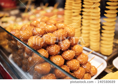 Stacked golden fried meatballs on skewers with sesame seeds displayed in a food stall, with spiral potato snacks visible in the background Stacked golden fried meatballs on skewers with sesame seeds displayed in a food stall, with spiral potato snacks visible in the background 140580689