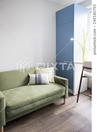 Green sofa with decorative pillows positioned beside a blue cabinet and a window, featuring a plant and a wooden chair in a modern interior setting 140580703