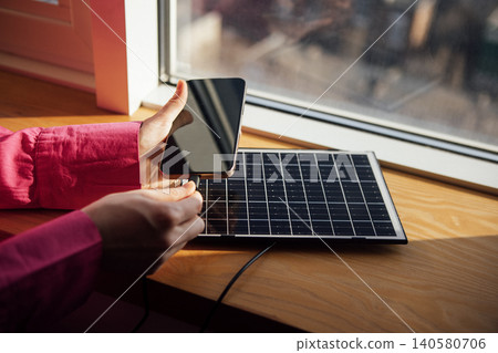 Person in pink shirt connects smartphone to small solar panel on wooden windowsill, with sunlight illuminating the scene and a blurred outdoor view in the background 140580706