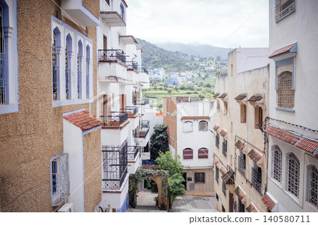 Residential buildings with traditional architecture lining a narrow street in a hillside town, showcasing white and beige facades with tiled roofs and distant mountains Residential buildings with traditional architecture lining a narrow street in a hillside town, showcasing white and beige facades with tiled roofs and distant mountains 140580711