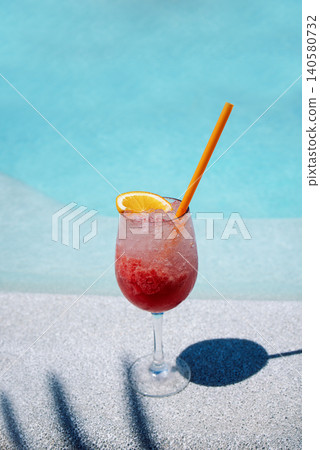 A delicious berry cocktail in an elegant glass on the side of the pool. A cup of cool summer drink with a slice of orange on a background of blue water. A delicious berry cocktail in an elegant glass on the side of the pool. A cup of cool summer drink with a slice of orange on a background of blue water. 140580732