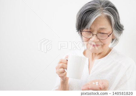 A gray-haired elderly woman (senior) taking medicine against a white background. An image representing health management, medication, medical care, and healthcare. A gray-haired elderly woman (senior) taking medicine against a white background. An image representing health management, medication, medical care, and healthcare. 140581820