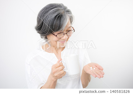 A gray-haired senior woman taking medicine in her hand against a white background. This image represents health management, medication, medical care, and healthcare. A gray-haired senior woman taking medicine in her hand against a white background. This image represents health management, medication, medical care, and healthcare. 140581826