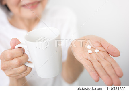 A gray-haired elderly woman (senior) taking medicine against a white background. An image representing health management, medication, medical care, and healthcare. A gray-haired elderly woman (senior) taking medicine against a white background. An image representing health management, medication, medical care, and healthcare. 140581831