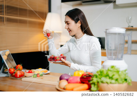 Healthy woman cooking at home while video calling online. Clean eating, nutrition, fresh vegetables, smoothie and digital lifestyle concept in modern kitchen. Healthy woman cooking at home while video calling online. Clean eating, nutrition, fresh vegetables, smoothie and digital lifestyle concept in modern kitchen. 140582682