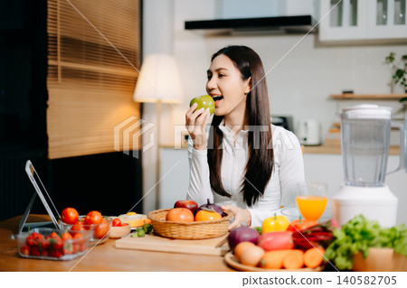 Smiling young woman preparing fresh strawberries, fruit and vegetables in a modern kitchen. Healthy lifestyle, clean eating, vegan nutrition and home cooking concept. 140582705