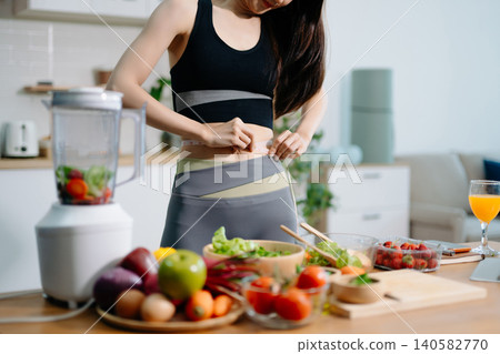 Woman measuring waist at home with fresh food. Healthy lifestyle, diet plan, weight loss, fitness, nutrition and wellness concept Woman measuring waist at home with fresh food. Healthy lifestyle, diet plan, weight loss, fitness, nutrition and wellness concept 140582770