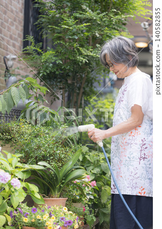 一位笑容可掬、頭髮花白的老婦人喜歡園藝,她正在為花園裡的花壇和植物澆水。 一位笑容可掬、頭髮花白的老婦人喜歡園藝,她正在為花園裡的花壇和植物澆水。 140582855
