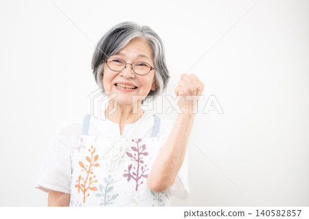 A senior woman with gray hair, wearing an apron, smiles at the camera and makes a triumphant gesture against a white background. A senior woman with gray hair, wearing an apron, smiles at the camera and makes a triumphant gesture against a white background. 140582857