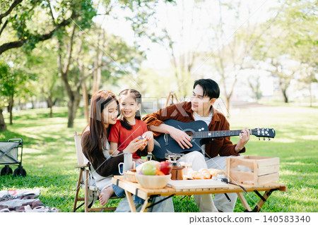 Joyful family enjoying a sunny picnic with music and laughter. Parents and child bonding outdoors, surrounded by love, warmth Joyful family enjoying a sunny picnic with music and laughter. Parents and child bonding outdoors, surrounded by love, warmth 140583340