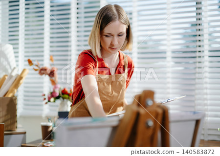 A focused female artist painting on canvas in a bright home studio, surrounded by natural light 140583872