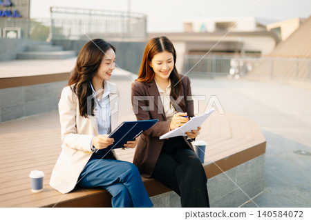 Asian businesswomen reviewing documents and writing notes outdoors. Modern teamwork, planning, and professional collaboration in a casual corporate lifestyle setting. Asian businesswomen reviewing documents and writing notes outdoors. Modern teamwork, planning, and professional collaboration in a casual corporate lifestyle setting. 140584072