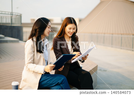 Asian businesswomen reviewing documents and writing notes outdoors. Modern teamwork, planning, and professional collaboration in a casual corporate lifestyle setting. Asian businesswomen reviewing documents and writing notes outdoors. Modern teamwork, planning, and professional collaboration in a casual corporate lifestyle setting. 140584073