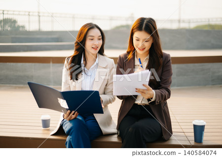 Asian businesswomen reviewing documents and writing notes outdoors. Modern teamwork, planning, and professional collaboration in a casual corporate lifestyle setting. Asian businesswomen reviewing documents and writing notes outdoors. Modern teamwork, planning, and professional collaboration in a casual corporate lifestyle setting. 140584074