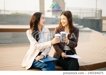 Asian businesswomen reviewing documents and writing notes outdoors. Modern teamwork, planning, and professional collaboration in a casual corporate lifestyle setting. Asian businesswomen reviewing documents and writing notes outdoors. Modern teamwork, planning, and professional collaboration in a casual corporate lifestyle setting. 140584075