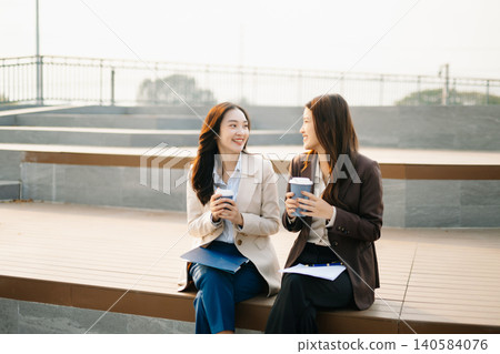 Asian businesswomen reviewing documents and writing notes outdoors. Modern teamwork, planning, and professional collaboration in a casual corporate lifestyle setting. Asian businesswomen reviewing documents and writing notes outdoors. Modern teamwork, planning, and professional collaboration in a casual corporate lifestyle setting. 140584076