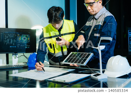 Engineers analyzing renewable energy project with wind turbine model and solar panels, reviewing digital blueprint on computer. 140585334