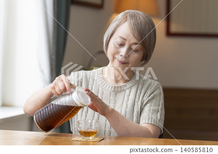 一位老婦人在客廳給自己倒了些大麥茶，然後喝了起來。 140585810