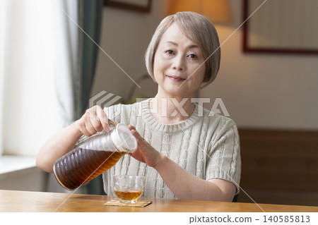 A senior woman pours herself some barley tea and drinks it in the living room. A senior woman pours herself some barley tea and drinks it in the living room. 140585813