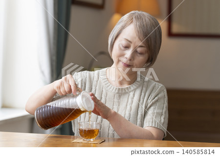 一位老婦人在客廳給自己倒了些大麥茶,然後喝了起來。 一位老婦人在客廳給自己倒了些大麥茶,然後喝了起來。 140585814