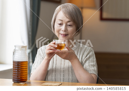 一位老婦人在客廳給自己倒了些大麥茶,然後喝了起來。 一位老婦人在客廳給自己倒了些大麥茶,然後喝了起來。 140585815