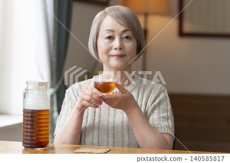 A senior woman pours herself some barley tea and drinks it in the living room. A senior woman pours herself some barley tea and drinks it in the living room. 140585817