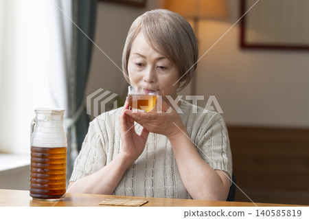 A senior woman pours herself some barley tea and drinks it in the living room. 140585819