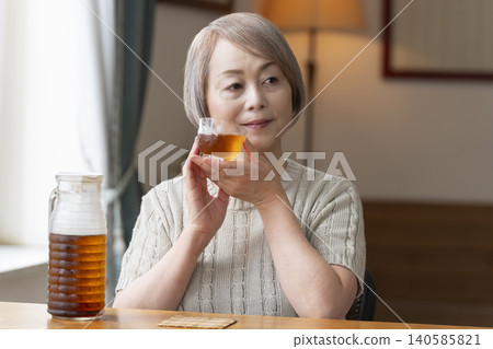 A senior woman pours herself some barley tea and drinks it in the living room. A senior woman pours herself some barley tea and drinks it in the living room. 140585821