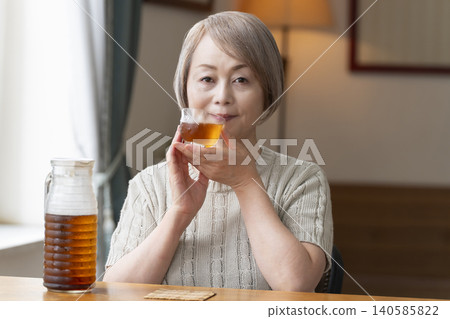 A senior woman pours herself some barley tea and drinks it in the living room. A senior woman pours herself some barley tea and drinks it in the living room. 140585822