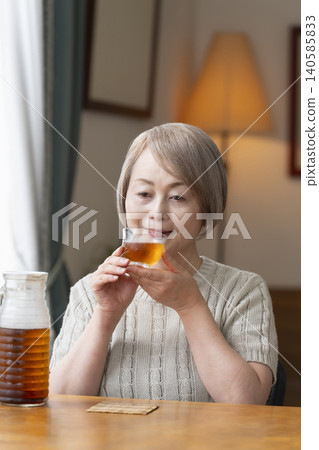 A senior woman pours herself some barley tea and drinks it in the living room. 140585833