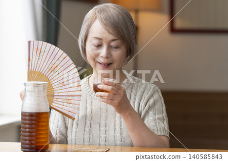 A senior woman pours herself some barley tea and drinks it in the living room. 140585843