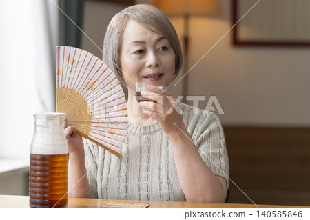 A senior woman pours herself some barley tea and drinks it in the living room. A senior woman pours herself some barley tea and drinks it in the living room. 140585846