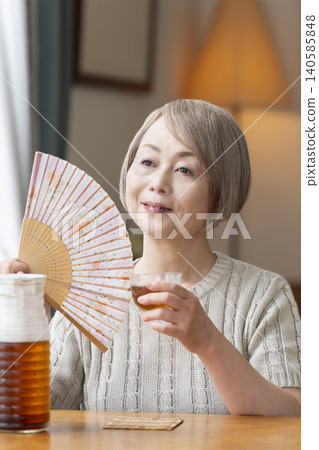 A senior woman pours herself some barley tea and drinks it in the living room. A senior woman pours herself some barley tea and drinks it in the living room. 140585848
