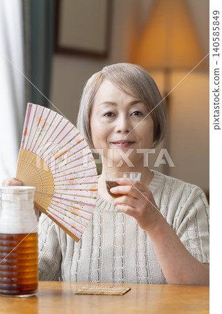 一位老婦人在客廳給自己倒了些大麥茶,然後喝了起來。 一位老婦人在客廳給自己倒了些大麥茶,然後喝了起來。 140585849