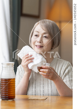 A senior woman pours herself some barley tea and drinks it in the living room. A senior woman pours herself some barley tea and drinks it in the living room. 140585852