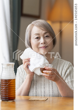 A senior woman pours herself some barley tea and drinks it in the living room. A senior woman pours herself some barley tea and drinks it in the living room. 140585853