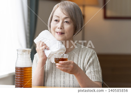 A senior woman pours herself some barley tea and drinks it in the living room. A senior woman pours herself some barley tea and drinks it in the living room. 140585856