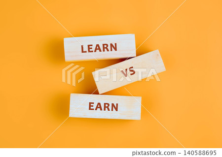 Three wooden blocks show the words learn and earn along with a small vs in the middle. The blocks sit against a bright orange surface Three wooden blocks show the words learn and earn along with a small vs in the middle. The blocks sit against a bright orange surface 140588695