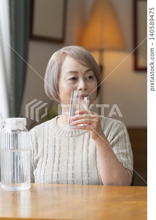 老年婦女在客廳喝水以預防中暑 老年婦女在客廳喝水以預防中暑 140589475