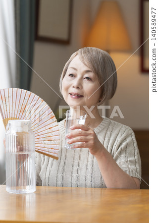 老年婦女在客廳喝水以預防中暑 老年婦女在客廳喝水以預防中暑 140589477