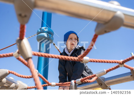 A young boy wearing a blue beanie framed by playground ropes at an outdoor adventure park. Candid lifestyle portrait capturing child concentration, outdoor activity, and active childhood. Cute little A young boy wearing a blue beanie framed by playground ropes at an outdoor adventure park. Candid lifestyle portrait capturing child concentration, outdoor activity, and active childhood. Cute little 140592356
